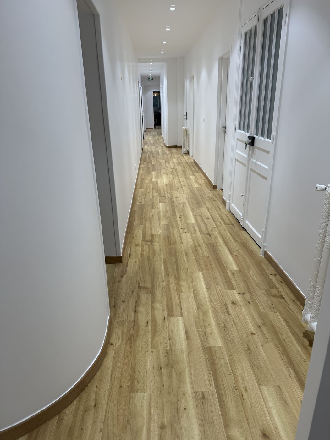 Long couloir blanc avec parquet clair, plinthes en bois et spots encastrés