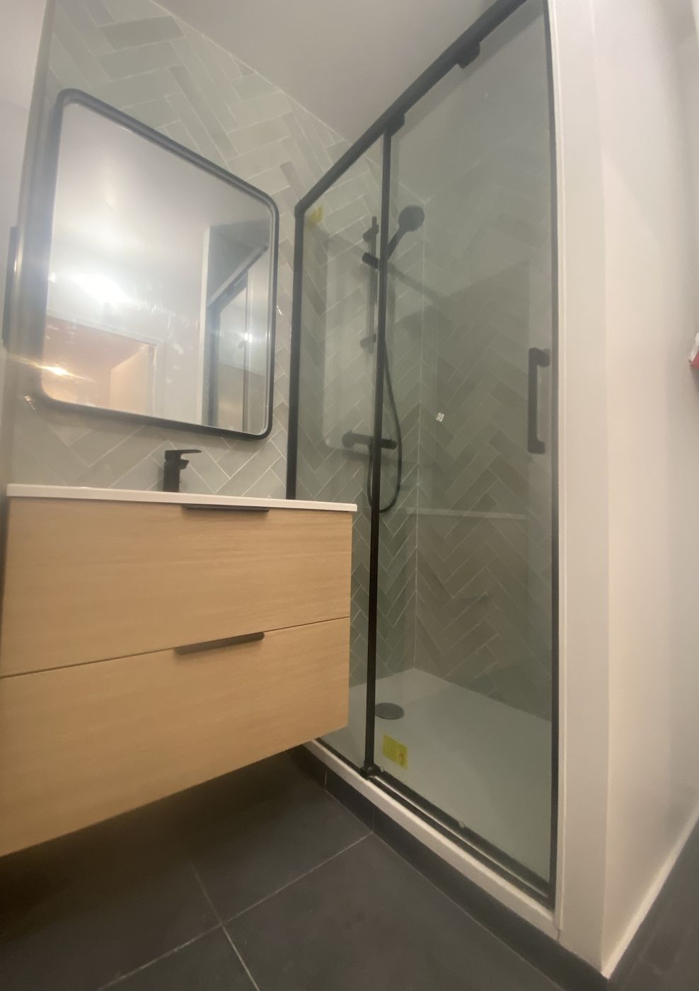 Salle de bain avec carrelage vert en chevrons, meuble en bois clair et douche vitrée noire